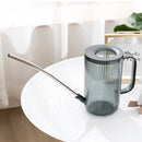 Watering can with extended spout on a table, surrounded by various tableware and drinkware items, primarily in grey tones.