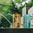Blue metal watering can with extended spout on grass, surrounded by wooden and glass objects.