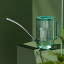 Stainless steel watering can with an extended spout on a table, featuring a transparent and metallic design for precise water delivery.