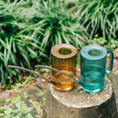 Metal watering can with an extended spout, placed on a wooden surface surrounded by plants and teacups.
