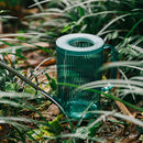 Green watering can with extended spout, surrounded by plants and grass in a natural garden environment.