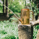 A metal watering can with an extended spout, positioned on wood near plants and grass, with trees in the background.