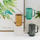 Watering can with extended spout on a wooden surface surrounded by grass and plants.