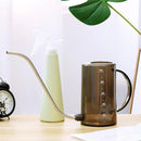 Watering tools set including a plastic watering can and a spray bottle arranged on a wooden table surrounded by plants.