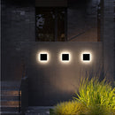 Waterproof LED outdoor wall lamp mounted on a brick building facade surrounded by plants, next to a window with a wooden rectangle design.