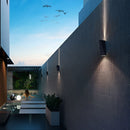 Modern Nordic style Empire waterproof LED outdoor wall light mounted on a building facade, with architectural elements and urban design features, including plants, a houseplant, and a shaded area under a sky with clouds and a bird visible.