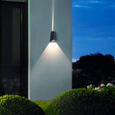 Outdoor wall-mounted pyramid LED sconce illuminating a building's exterior with plants, trees, and grass under a clear sky.