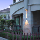 Outdoor pyramid LED wall sconce mounted on a house next to a window, surrounded by plants and a fence, showcasing its waterproof and lighting features.