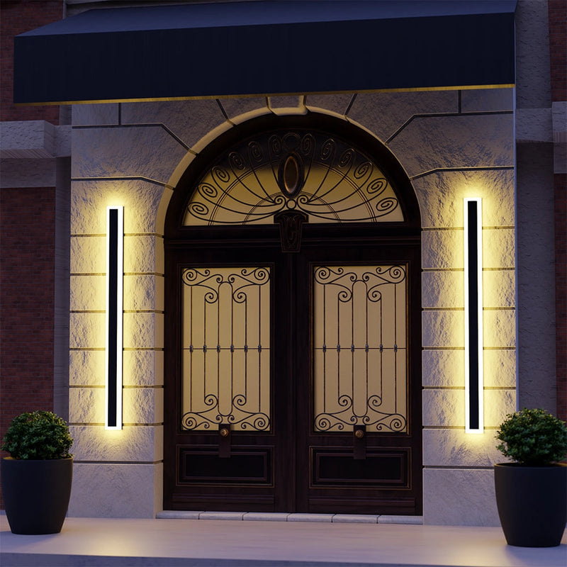 Minimalist LED wall sconce with sleek vertical rectangular design, mounted on a modern interior wall near a wooden door and a flowerpot with houseplants. The light is 32W, dimmable, and features a motion detector.