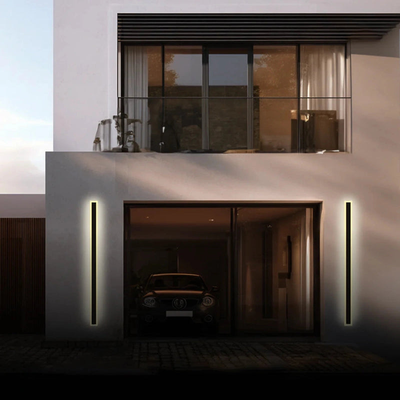 A sleek vertical rectangular minimalist LED wall sconce mounted on an exterior building wall near a window and door.