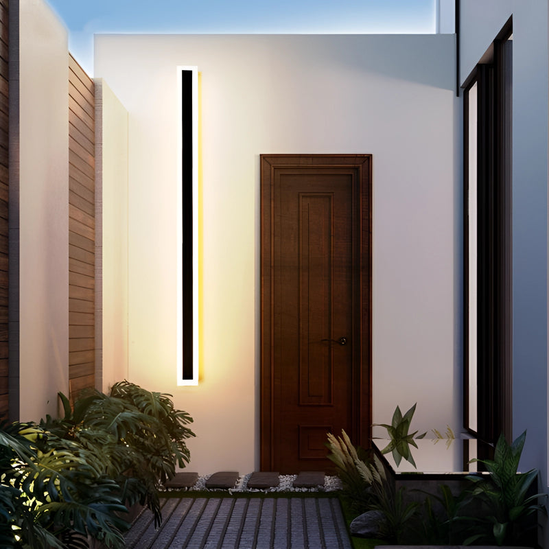 A sleek vertical rectangular LED wall sconce with a minimalist design, mounted on a wooden wall, next to a door and a potted plant, illuminating the interior space.