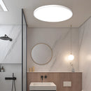 Minimalist round LED ceiling light in white, flush mounted on a ceiling. The slim, energy-efficient fixture illuminates a modern bathroom with white cabinetry, a mirror, and a sink.