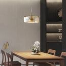 Modern glass pendant light with brass accents hanging from the ceiling, illuminating an elegantly designed interior with wooden furniture, a table, chairs, and decorative plants.
