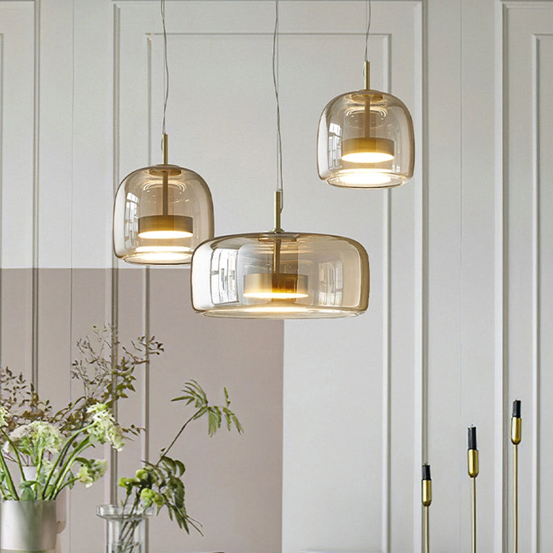 Modern glass pendant light with brass accents hanging from the ceiling, illuminating a stylish interior space with white walls, wooden furniture, and a houseplant in the background.