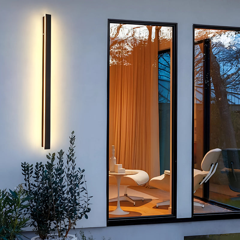 Slim Vertical LED Wall Sconce illuminated on a wall with a minimalist design, surrounded by modern interior decor including a plant, wooden elements, and a door.