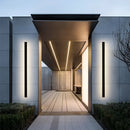 Slim vertical LED wall sconce with minimalist design on a building exterior, surrounded by plants and modern architecture.