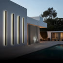 Slim Vertical LED Wall Sconce mounted on a modern building facade with a minimalist design, illuminated against a backdrop of sky and trees.
